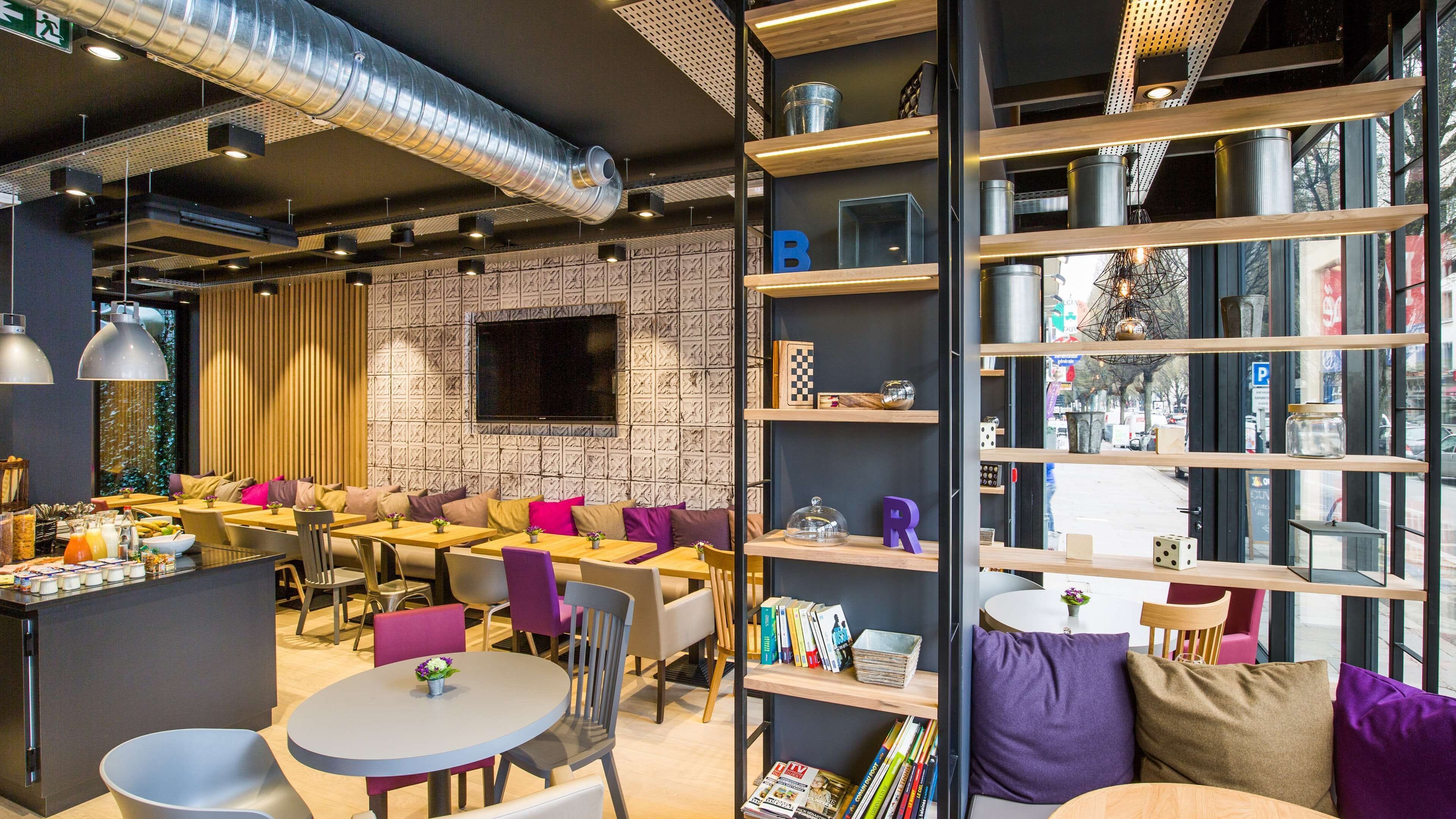 Ein modernes Café mit runden Tischen, violetten Stühlen und einem Bücherregal mit verschiedenen Gegenständen.