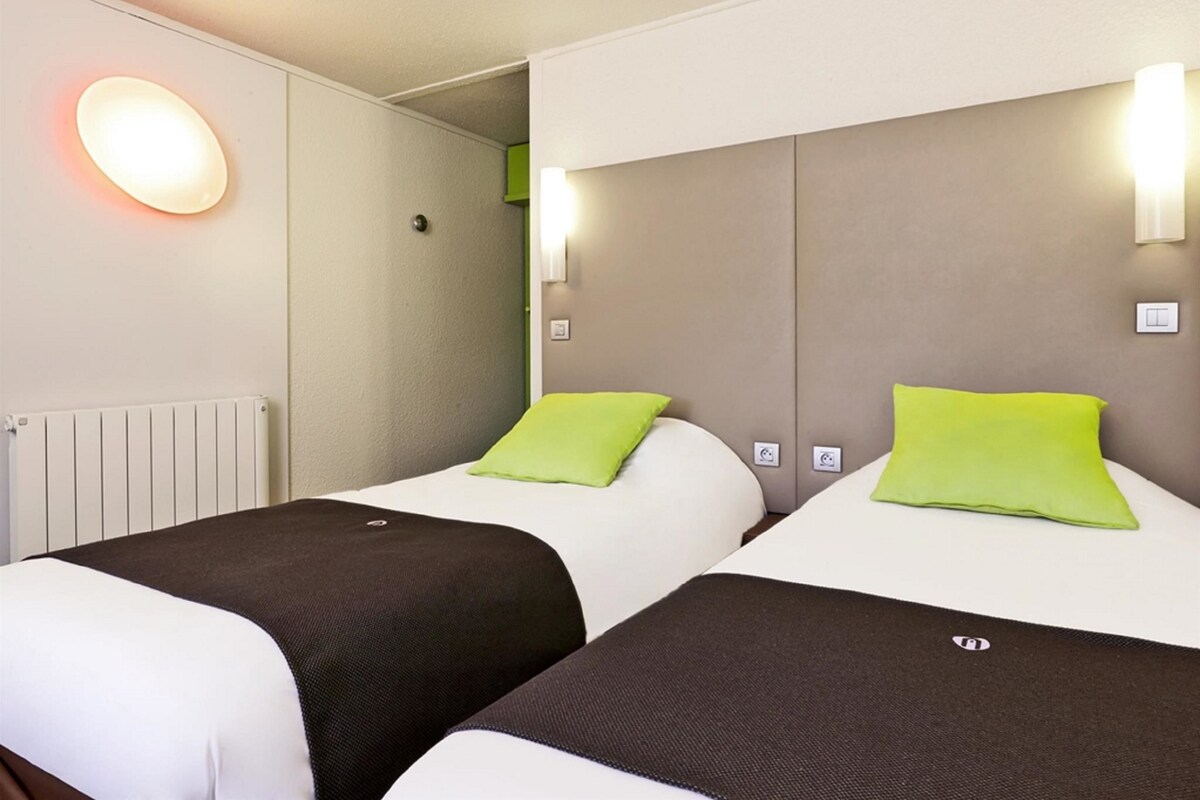 Habitación de hotel con dos camas, cada una con almohada verde y cabecera blanca. Se observan luces fijadas a la pared y un enchufe visible.