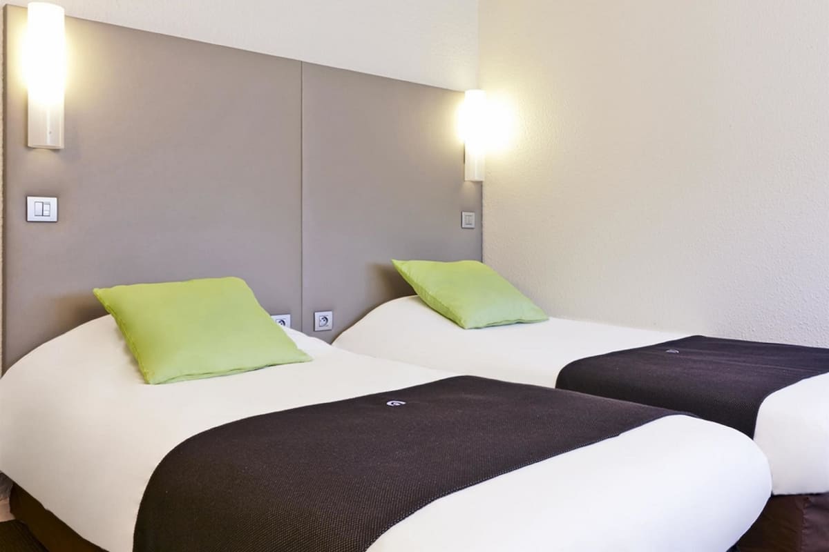 Une chambre d’hôtel avec deux lits, chacun agrémenté d’un oreiller vert et d’une couverture noire et blanche. Des appliques murales sont fixées de part et d’autre des lits.