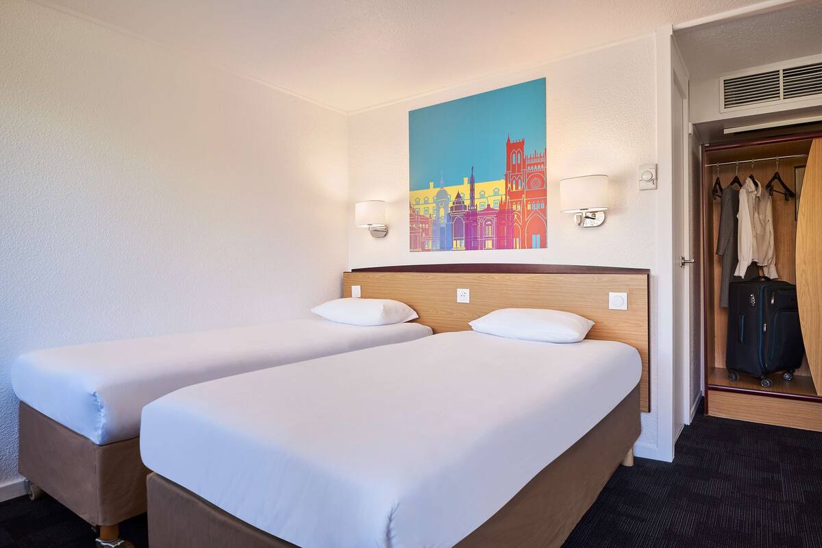 A hotel room with two single beds, a wooden headboard, a colorful cityscape painting, a wardrobe, and a suitcase.
