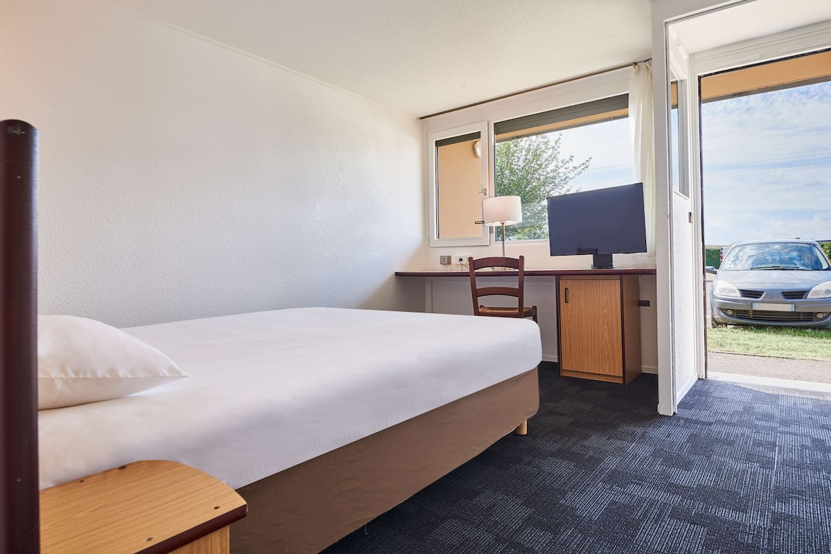 A hotel room with a bed, a desk, a chair, a TV, and a view of a car through the door.