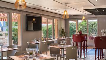 A restaurant with multiple tables and chairs, large windows with views of greenery, and hanging pendant lights.