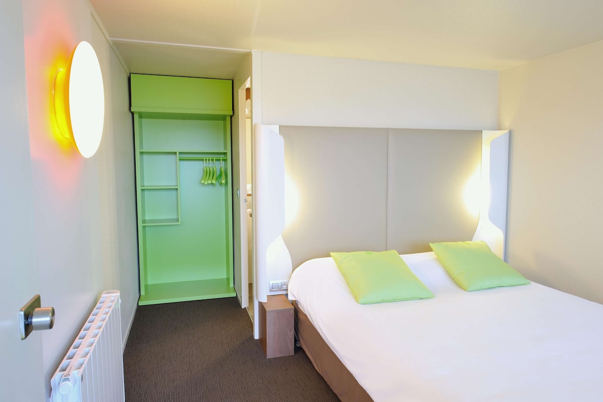 Una habitación de hotel con una cama, un armario verde, un radiador y una lámpara fijada a la pared.