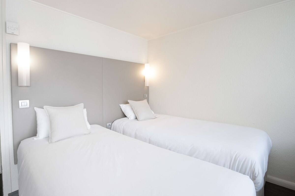 A hotel room with two single beds, each with white bedding and pillows. There are wall-mounted light fixtures on either side of the beds.
