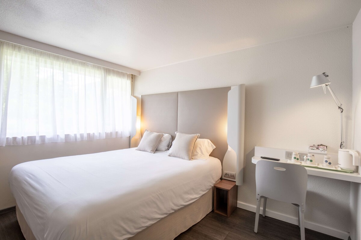 A hotel room with a large bed, a desk with a chair, a lamp, and a nightstand.