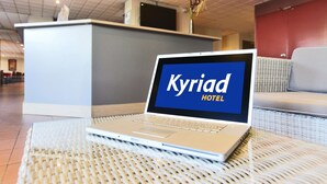 Een laptop op een rieten tafel waarop 'Kyriad Hotel' staat, met een receptiebalie en zithoek op de achtergrond.