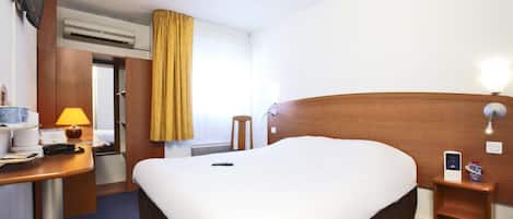 A hotel room with a large bed, a desk with a lamp, a TV mounted on the wall, and a bedside table with a phone and a small box.