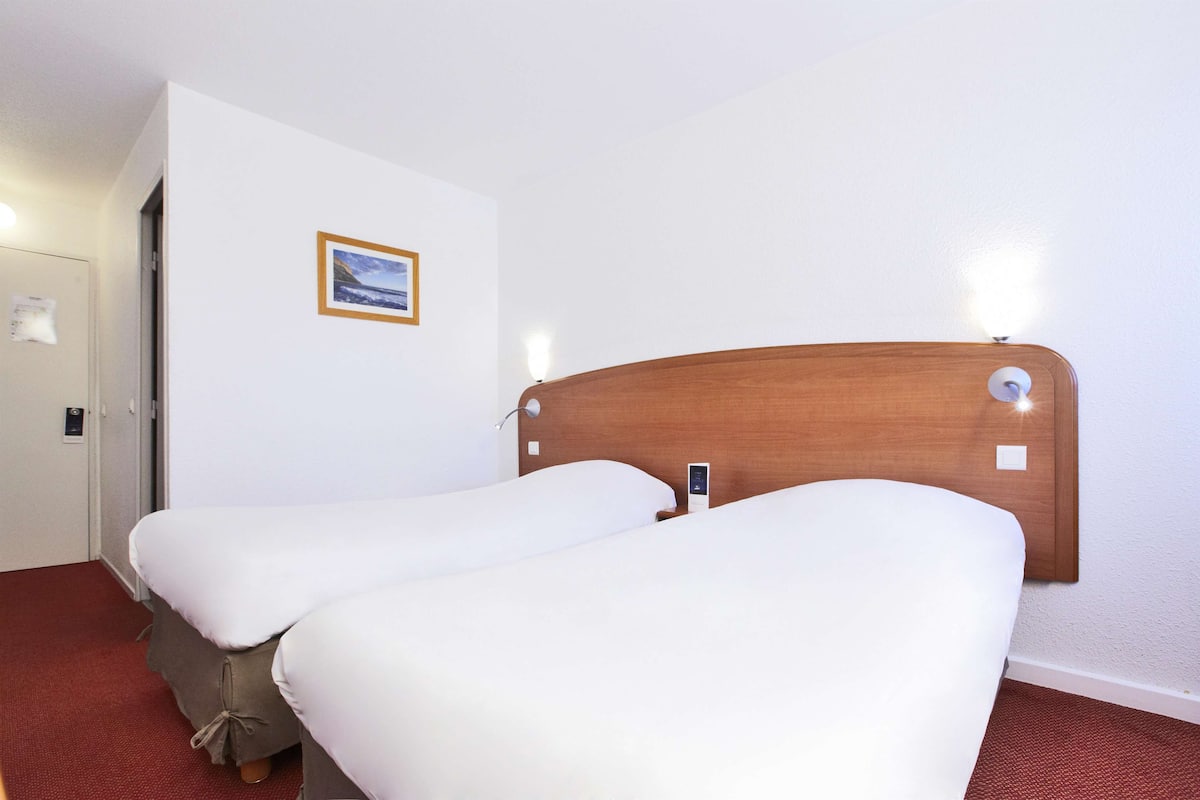 Une chambre d’hôtel avec deux lits simples, une tête de lit en bois, une petite table de chevet, une commode, une lampe fixée au mur et un tableau encadré accroché au mur.