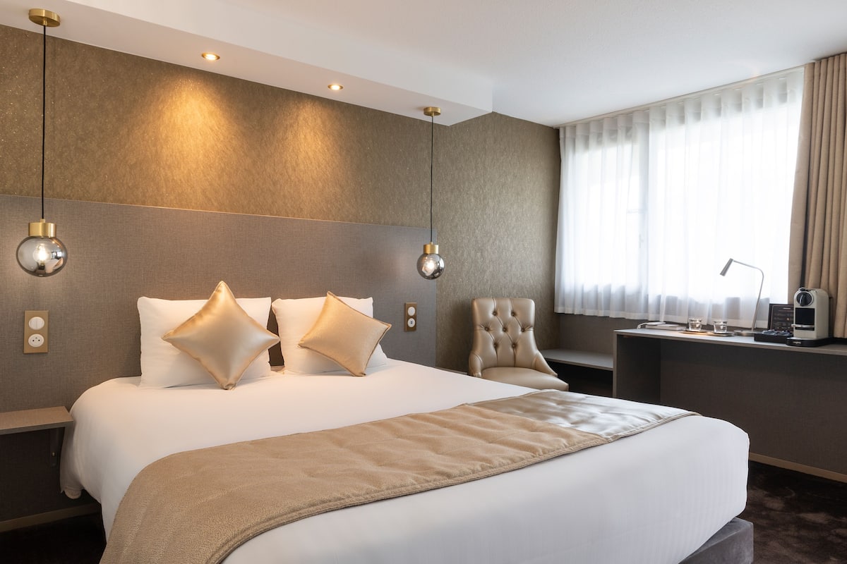 Ein modernes Hotelzimmer mit einem großen Bett, zwei Nachttischen, einem Schreibtisch mit Stuhl und einem großen Fenster mit Vorhängen.