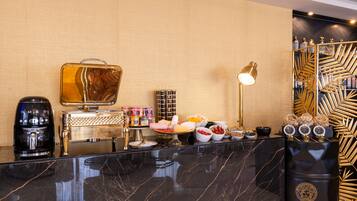 Un espace d’accueil d’hôtel avec un comptoir en marbre, une machine à café, un buffet proposant divers plats et une composition florale décorative.