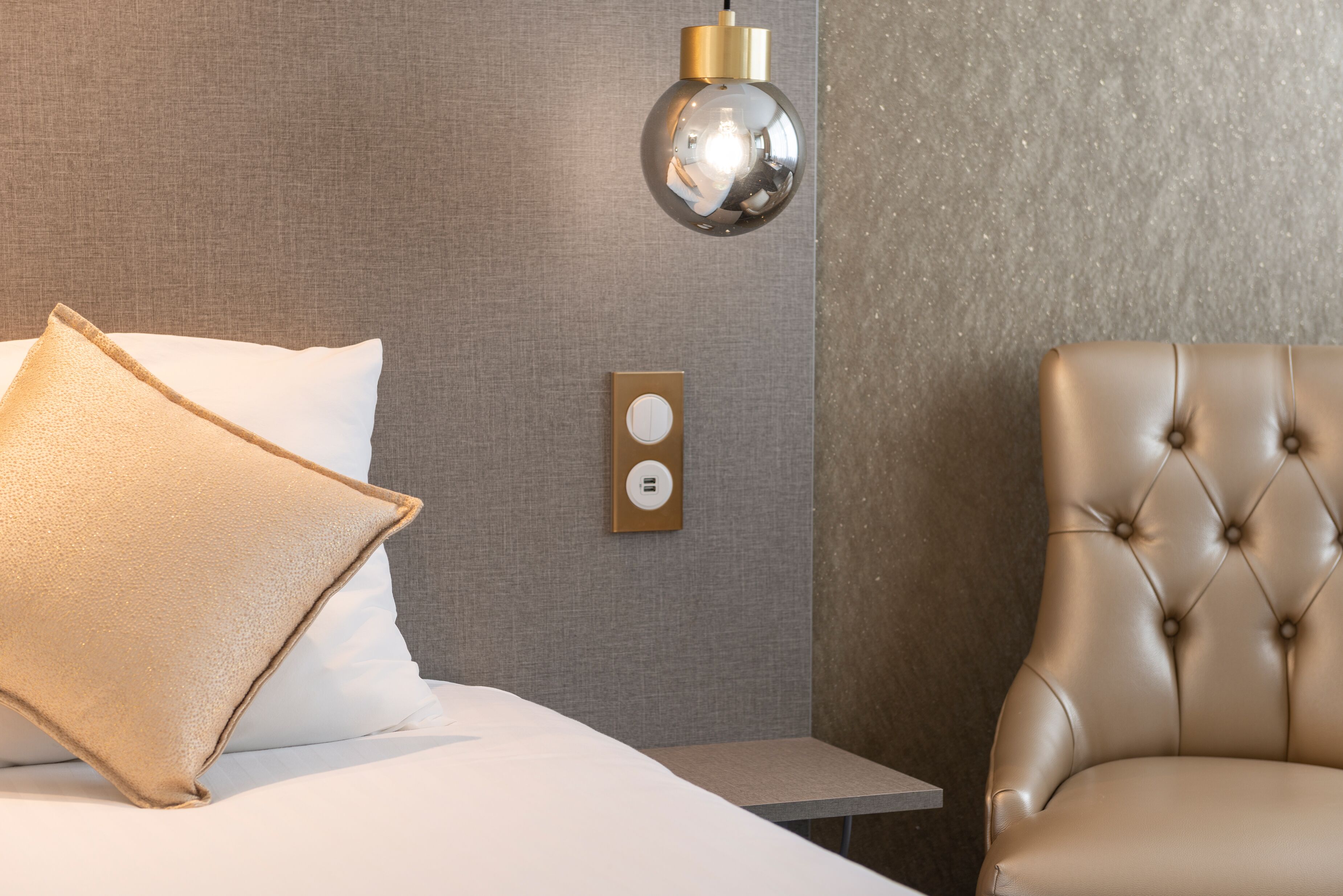 A hotel room with a bed, a chair, a wall-mounted light fixture, and a power outlet.