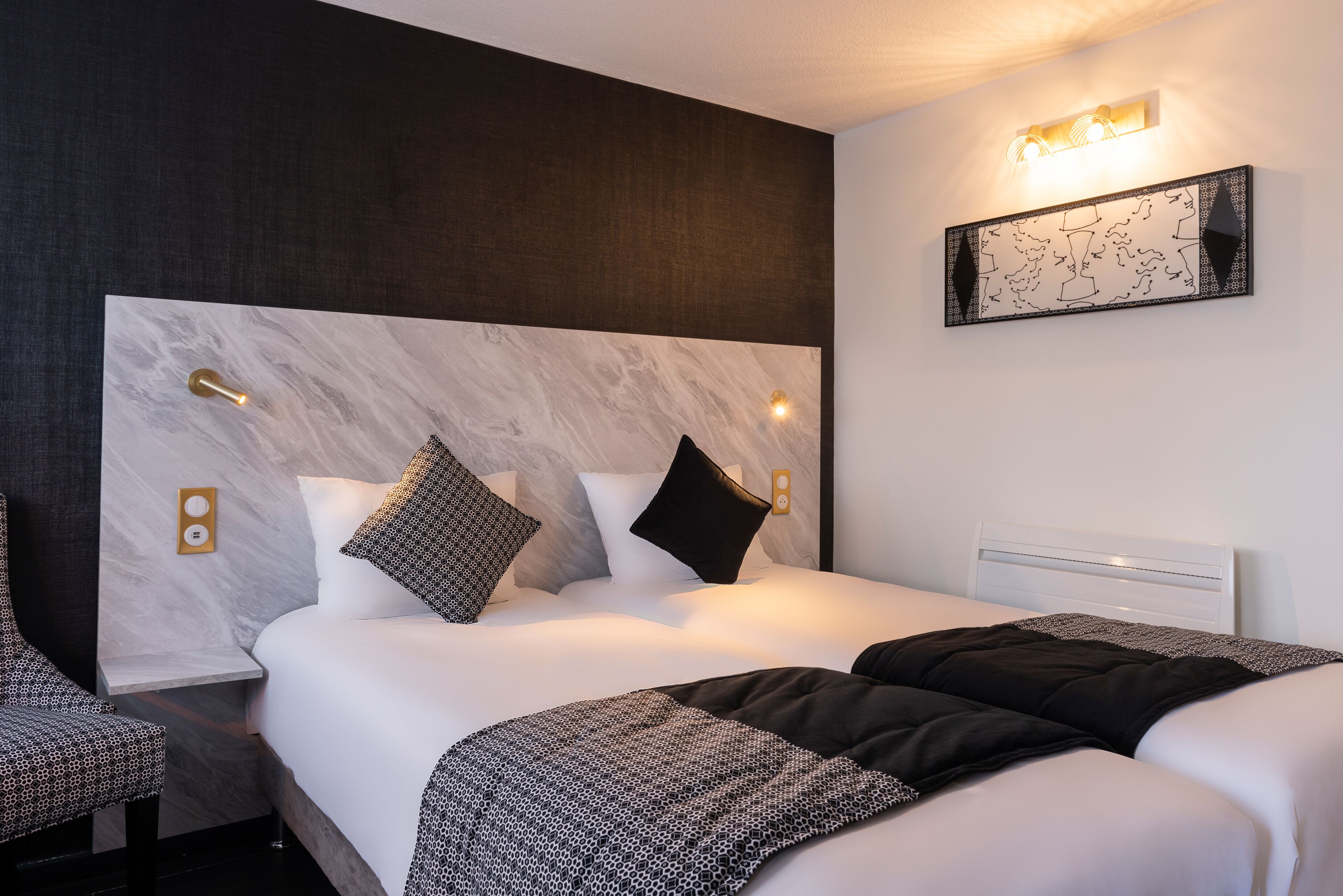 A modern hotel room with a marble headboard, a bed with black and white bedding, a bedside table, and a wall-mounted light fixture.
