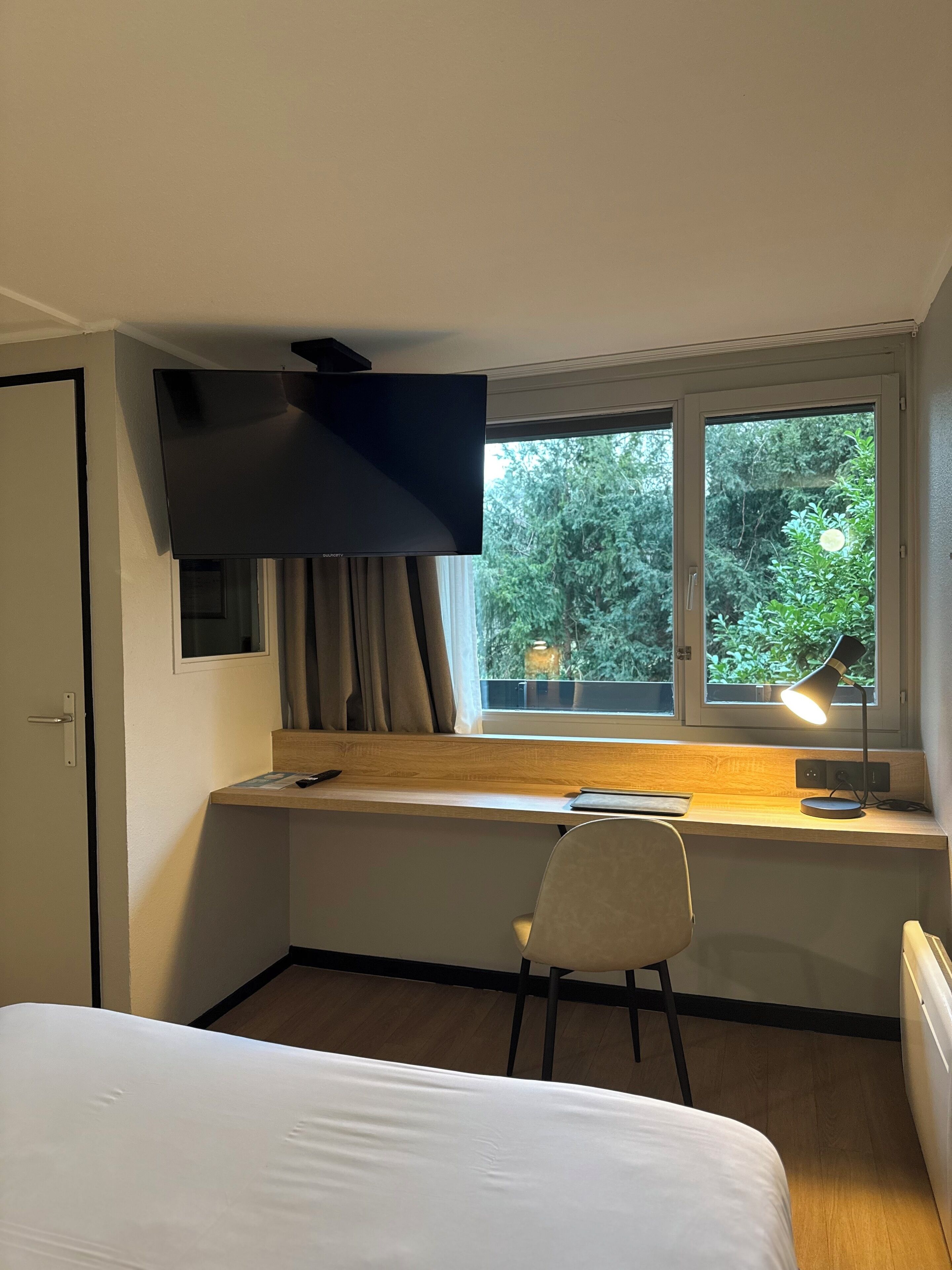 Habitación de hotel con cama, escritorio con silla, televisor montado en la pared y ventana con vista a zonas verdes.