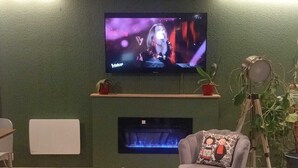 A modern living room with a flat-screen TV mounted on a green wall, a fireplace below, and contemporary furniture including a grey armchair and a black chair. - Campanile Nature Haguenau (Haguenau)
