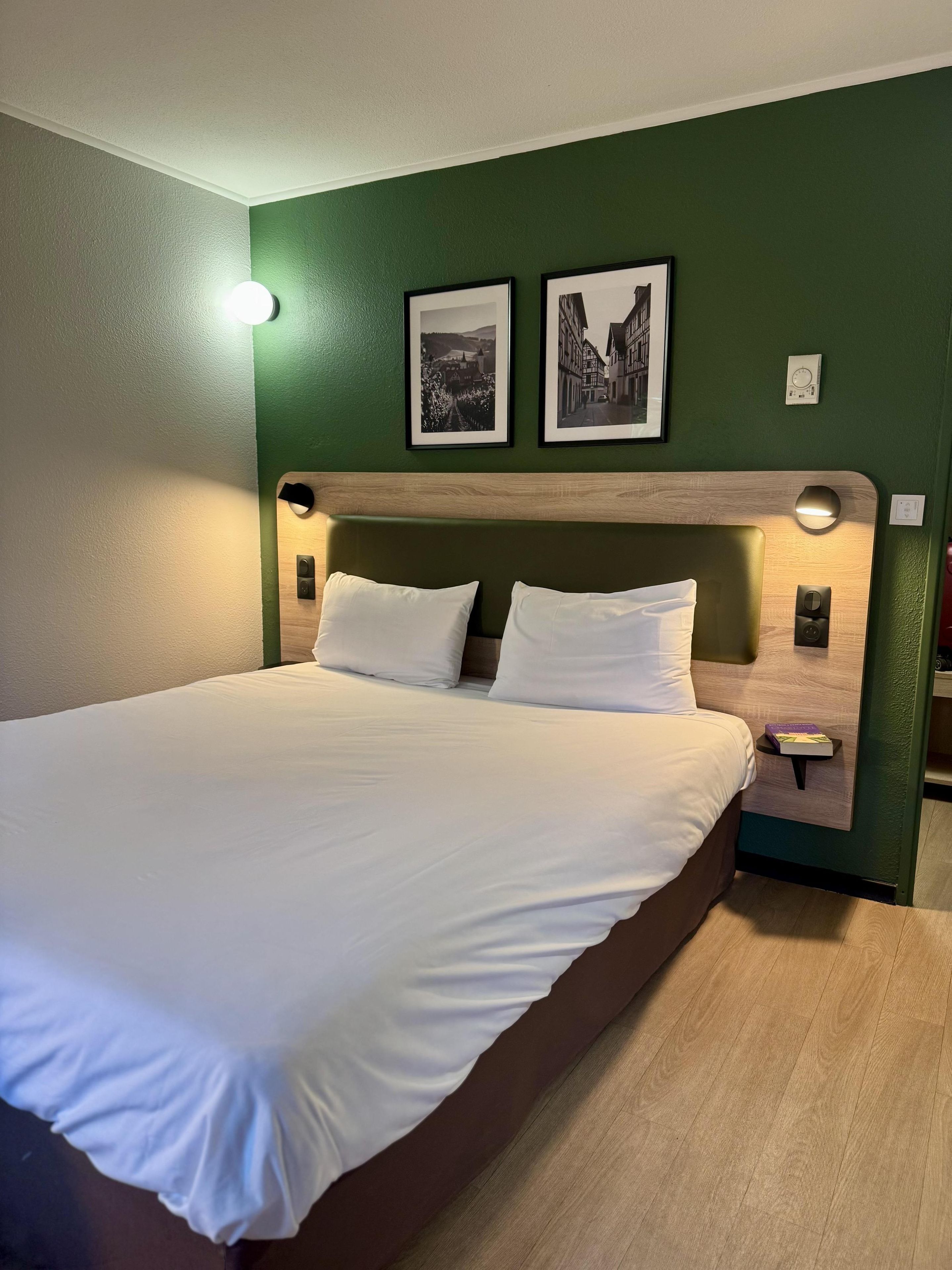 Un lit bien fait dans une pièce aux murs verts, avec deux tableaux encadrés et une tête de lit en bois.