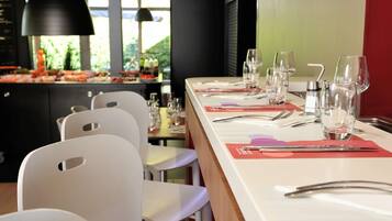 A restaurant with a long dining table set for a meal, white chairs, and a view of the outdoors through large windows.