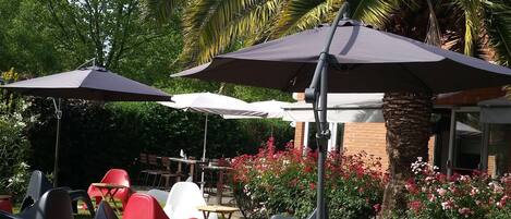An outdoor seating area with plastic chairs and umbrellas, surrounded by greenery and a brick building.