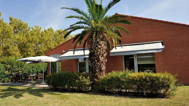 A building with a red brick exterior, a palm tree, and a covered outdoor seating area. - Campanile Toulouse Sud - Labege Innopole (Labege)
