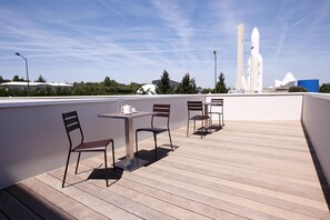 Eine Dachterrasse mit Tisch und Stühlen, im Hintergrund ist eine Raketenstartanlage sichtbar.