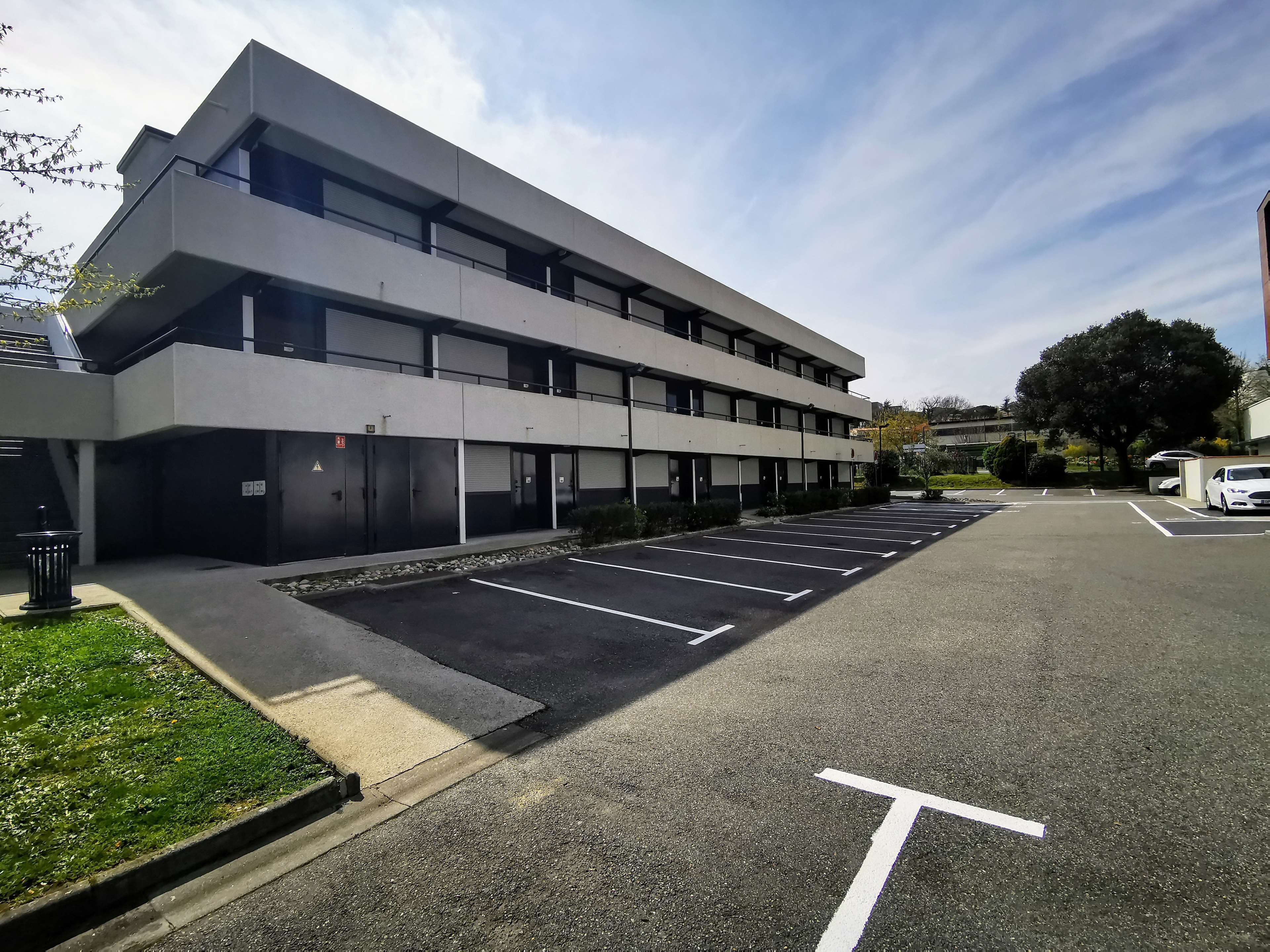 A modern building with a parking lot, multiple floors, and a large tree in the background.