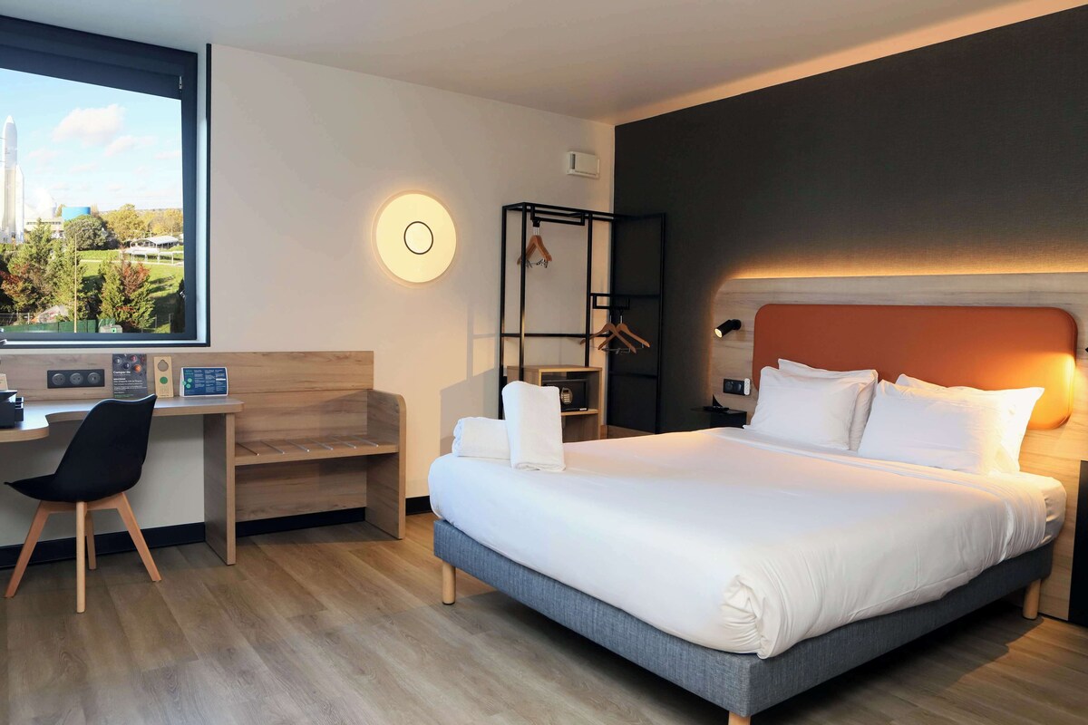 Une chambre d’hôtel moderne avec un grand lit, un bureau et une chaise. La fenêtre offre une vue sur le lancement d’une fusée.