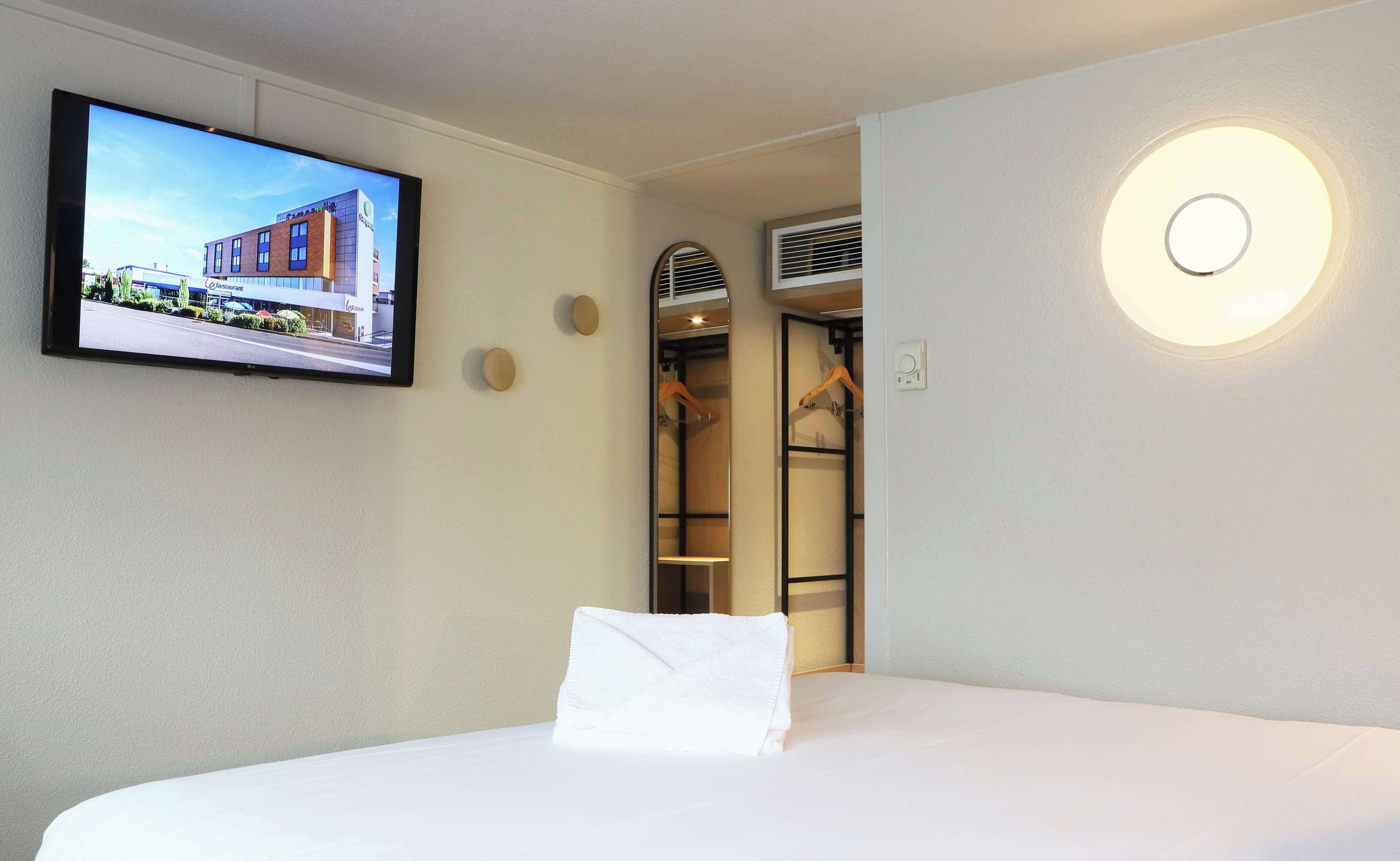 A hotel room with a bed, a television mounted on the wall, a closet with hanging clothes, and a circular light fixture.