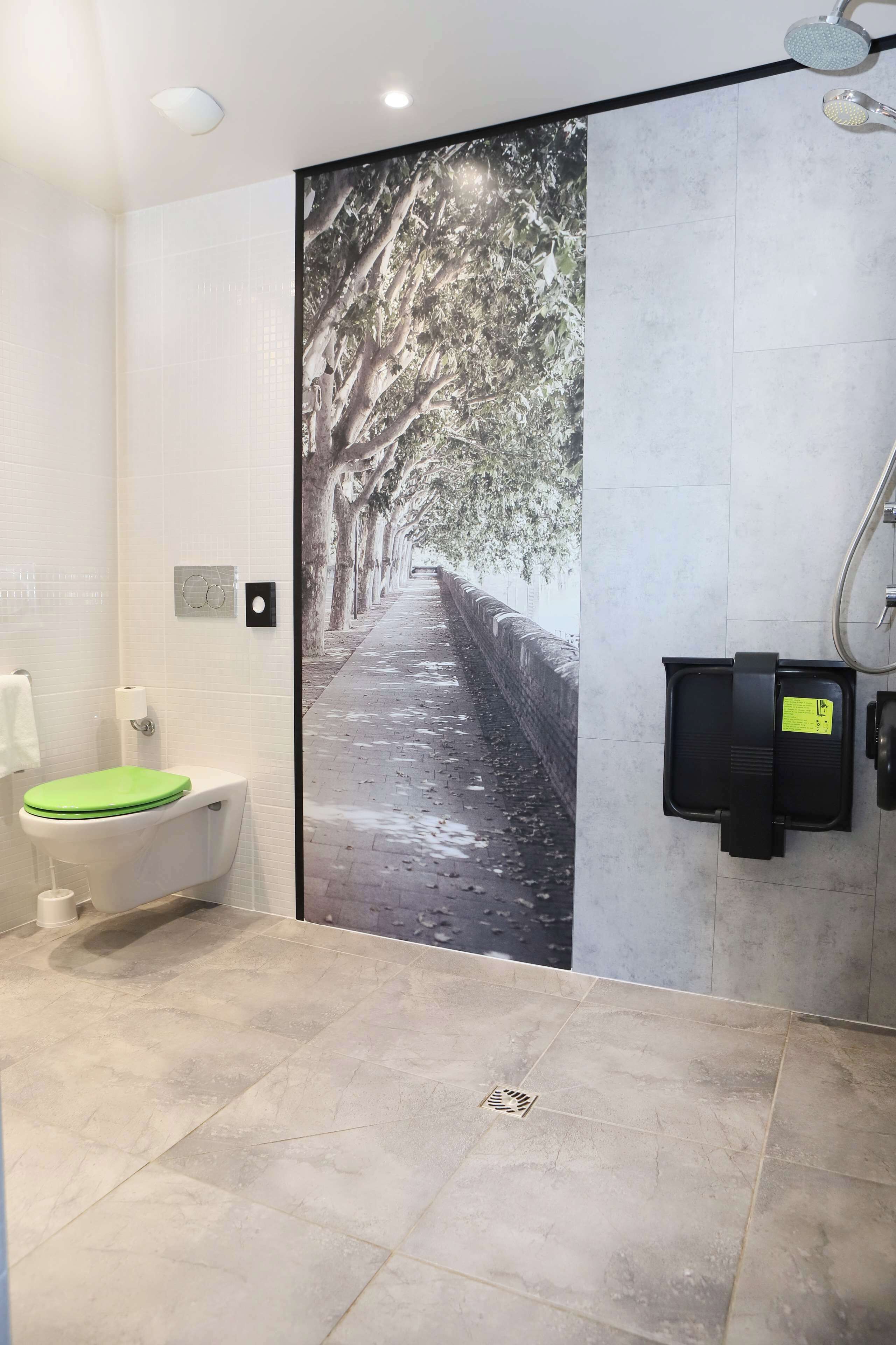 Un baño con un gran mural en la pared que muestra un sendero bordeado de árboles, un inodoro blanco con asiento verde, y una zona de ducha con cabezal de ducha manual.