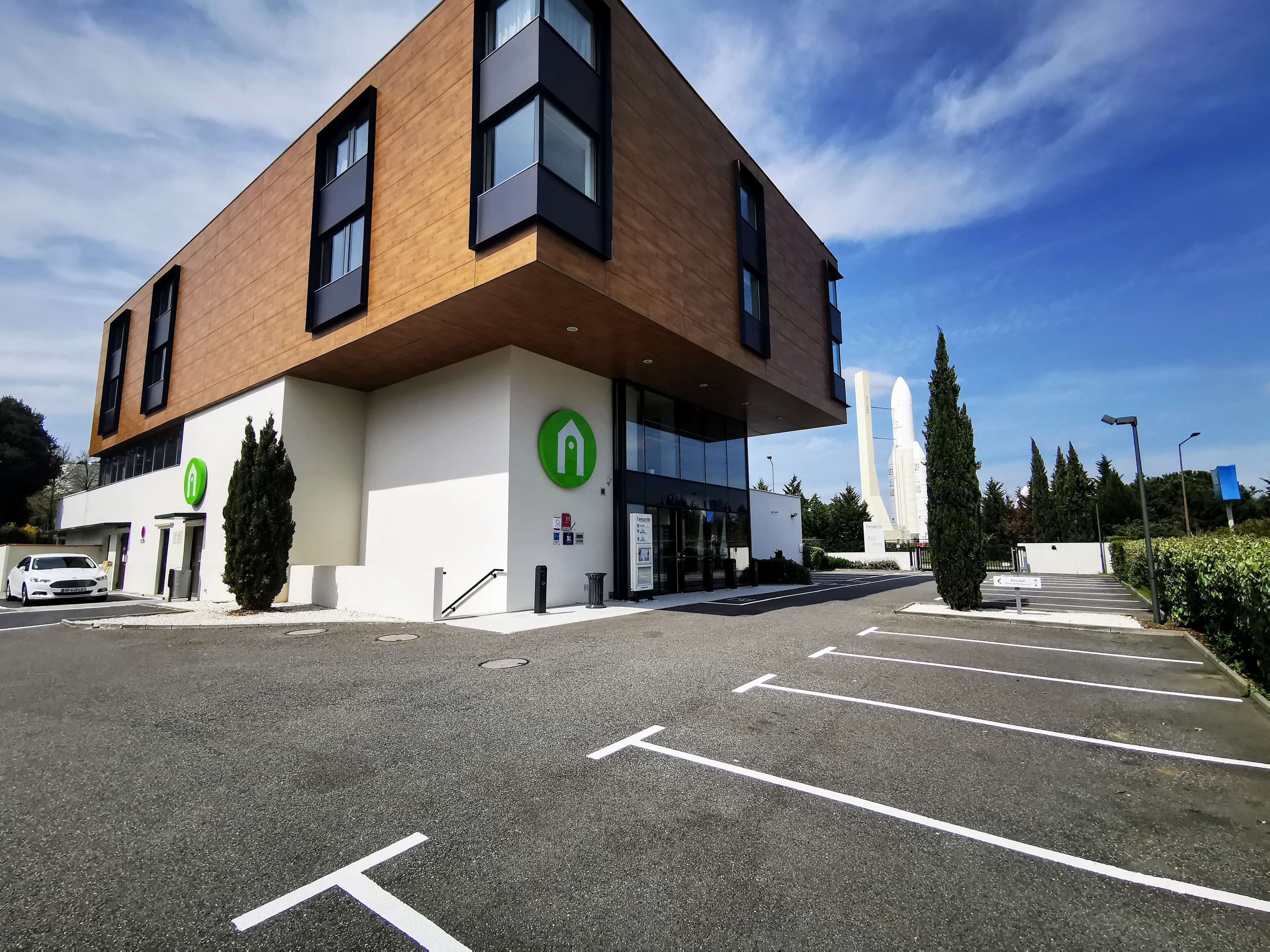 A modern building with a parking lot, a green logo, and a tall structure in the background.