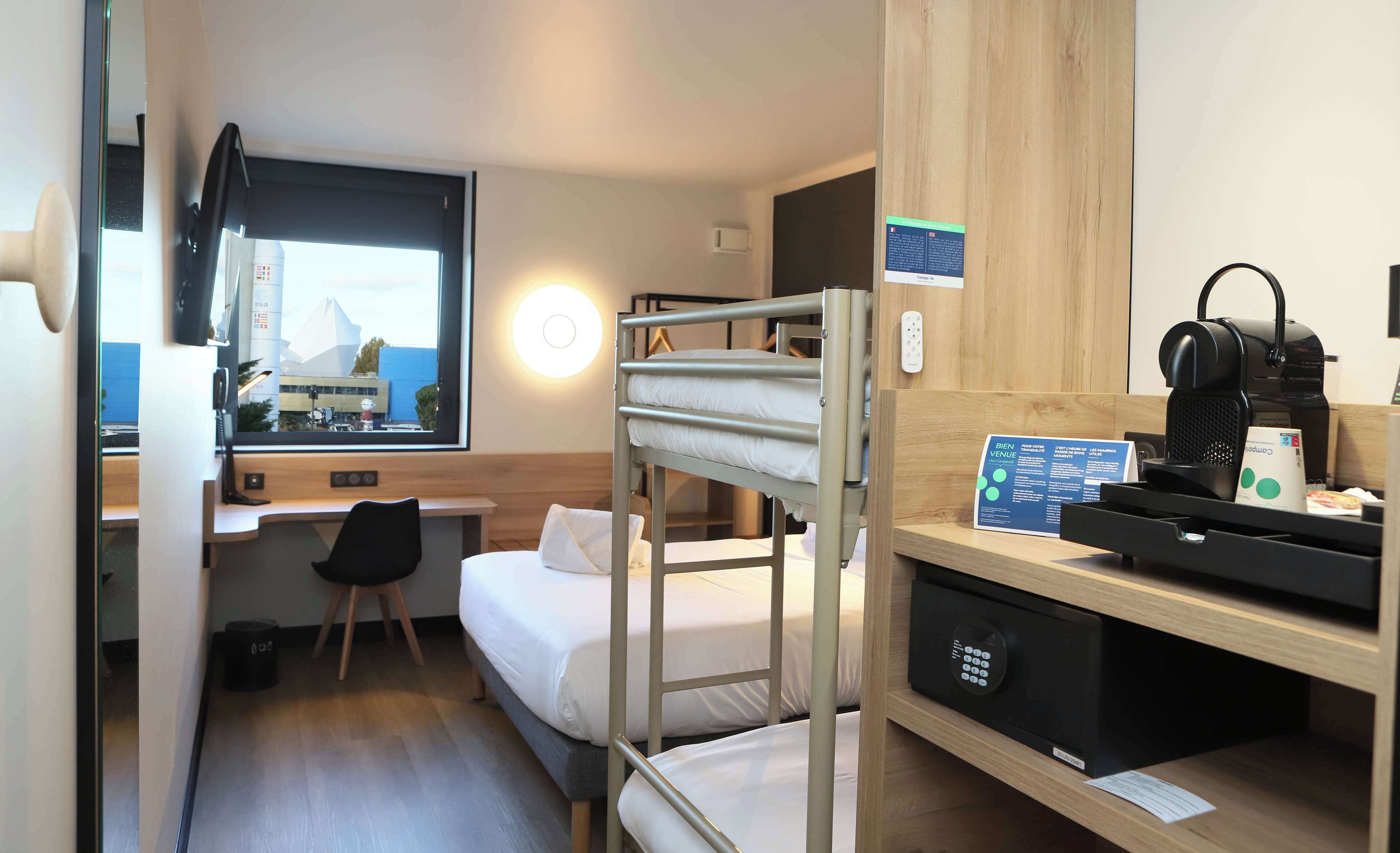 Habitación de hotel compacta con literas, un escritorio con computadora, una silla, una máquina de café y vistas a un edificio a través de una ventana.