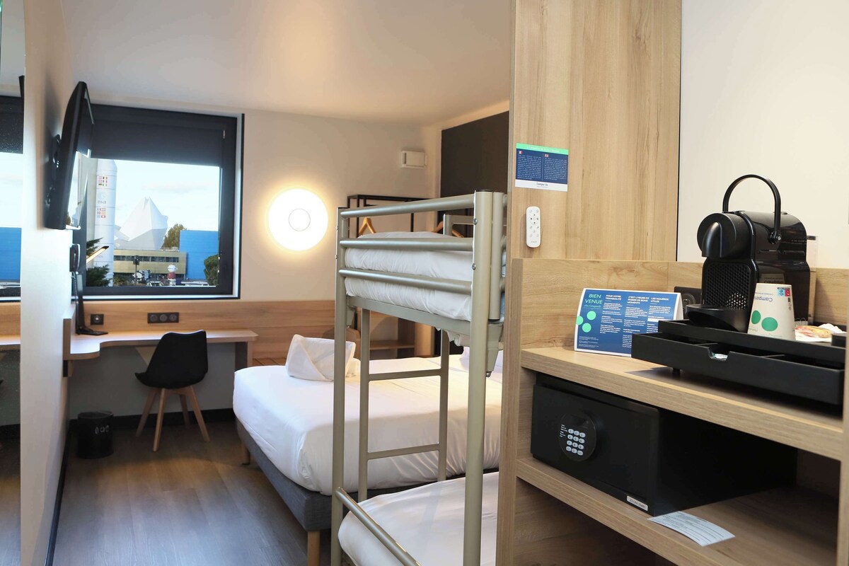 Une chambre d’hôtel compacte avec des lits superposés, un bureau avec un ordinateur, une chaise, une machine à café et une vue sur un immeuble par la fenêtre.