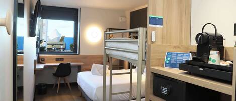 Habitación de hotel compacta con literas, un escritorio con computadora, una silla, una máquina de café y vistas a un edificio a través de una ventana.