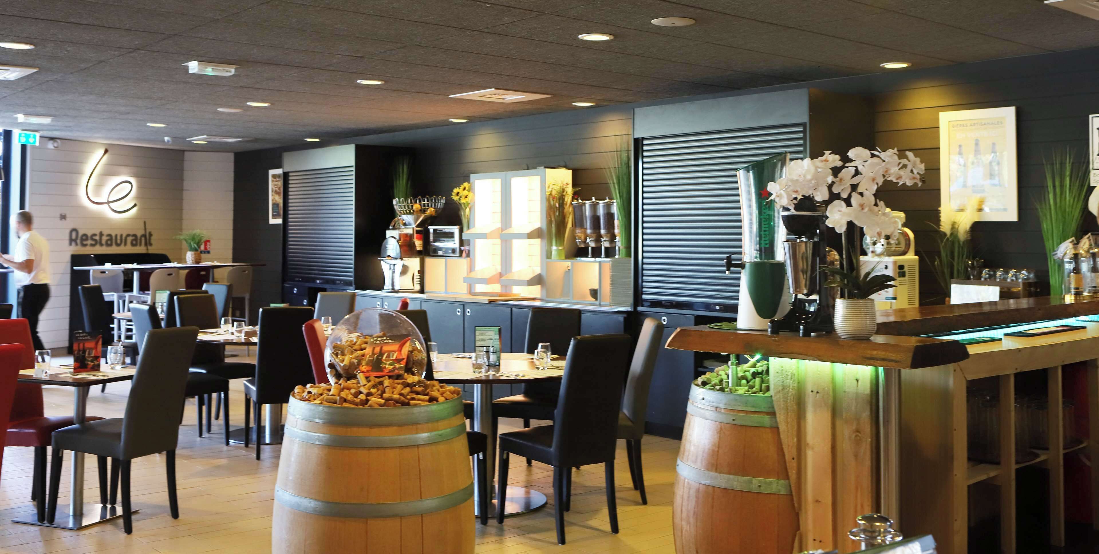 A restaurant interior with wooden barrels, tables, chairs, a bar area, and decorative plants.