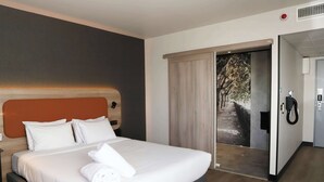 A hotel room with a large bed, a wooden headboard, and a view of a tree-lined path through an open door. - Campanile PRIME - Toulouse Sud Balma Cité de l'Espace (Toulouse)