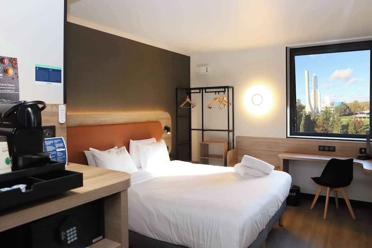 A hotel room with a bed, a desk, a coffee machine, and a window with a view of a rocket launch.