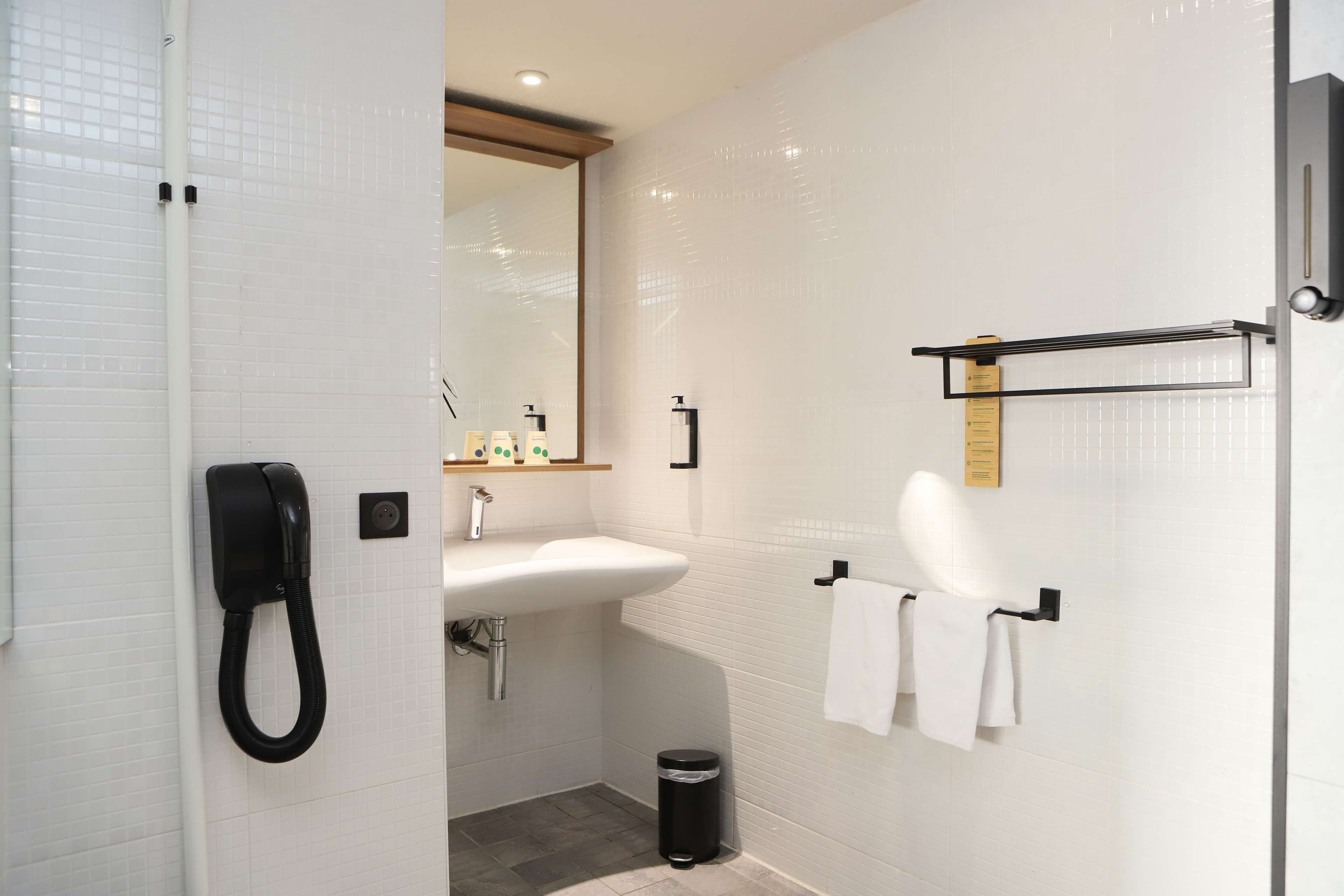 Casa de banho com lavatório branco, espelho, porta-toalhas e um caixote do lixo preto.