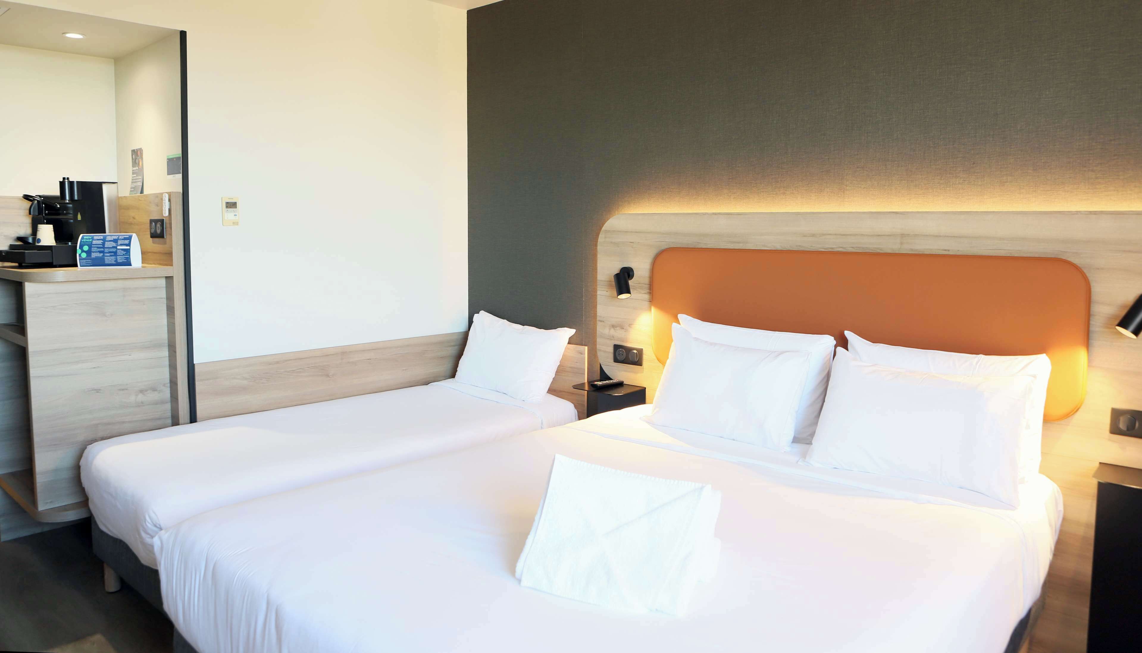 A hotel room with two beds, a desk, a coffee machine, and a wall-mounted lamp.