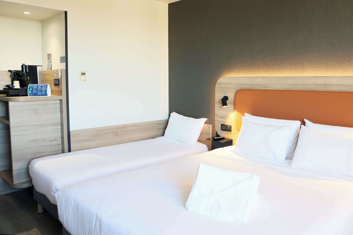 Une chambre d’hôtel avec deux lits, un bureau, une machine à café et une lampe fixée au mur.