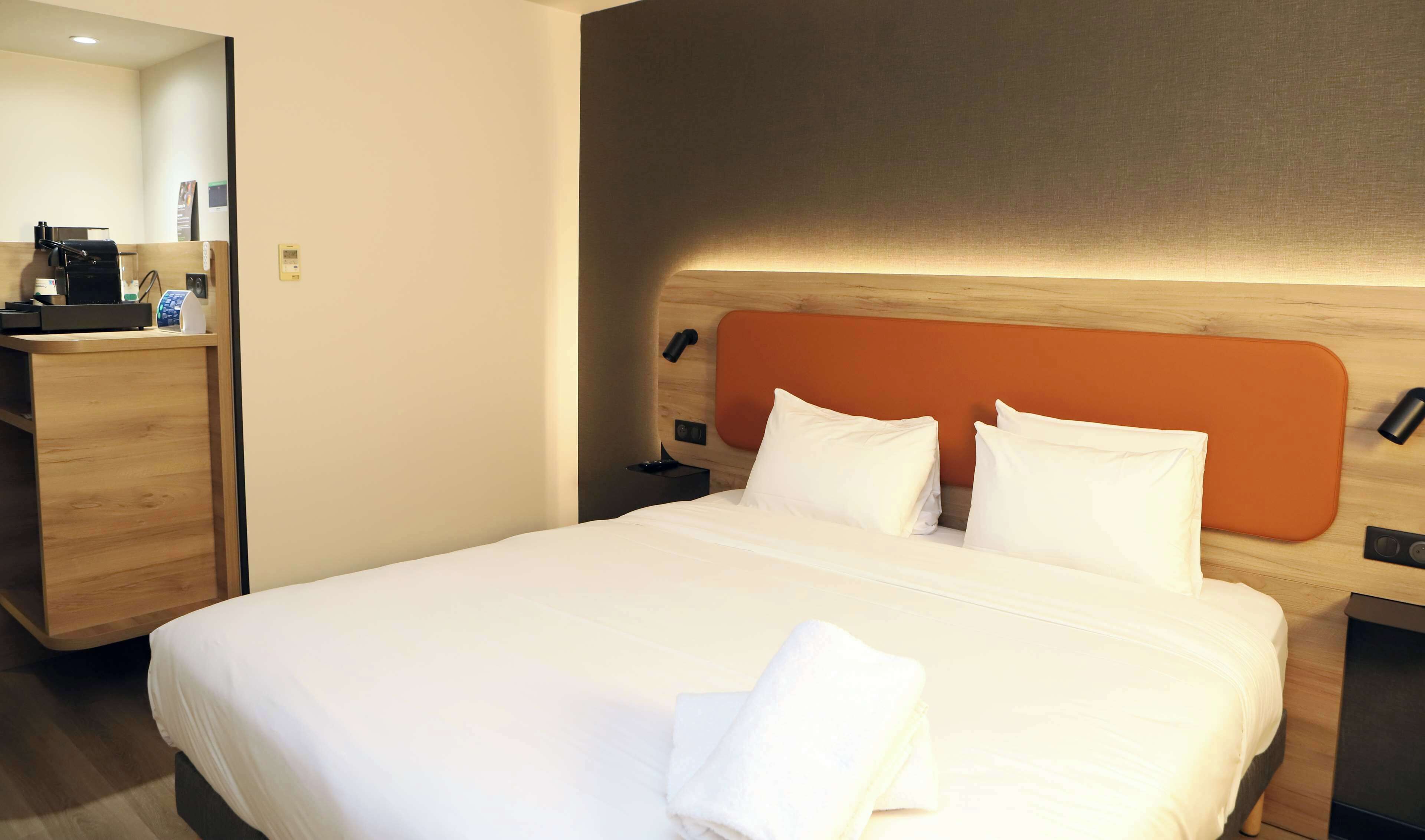 A hotel room with a neatly made bed, a wooden desk with a coffee machine, and a modern headboard.