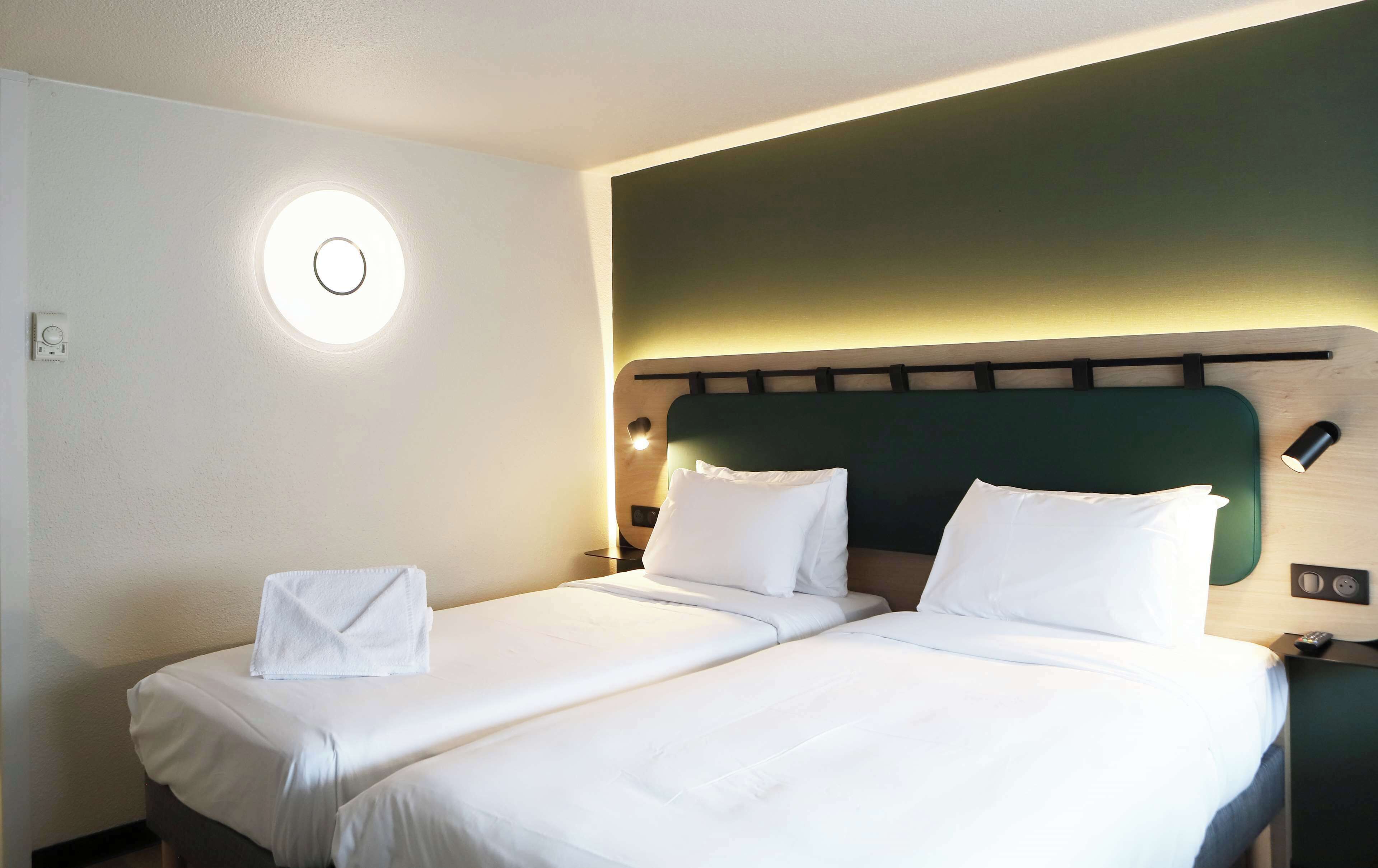 A hotel room with a bed, a round wall light, a thermostat, and a bedside table with a lamp.