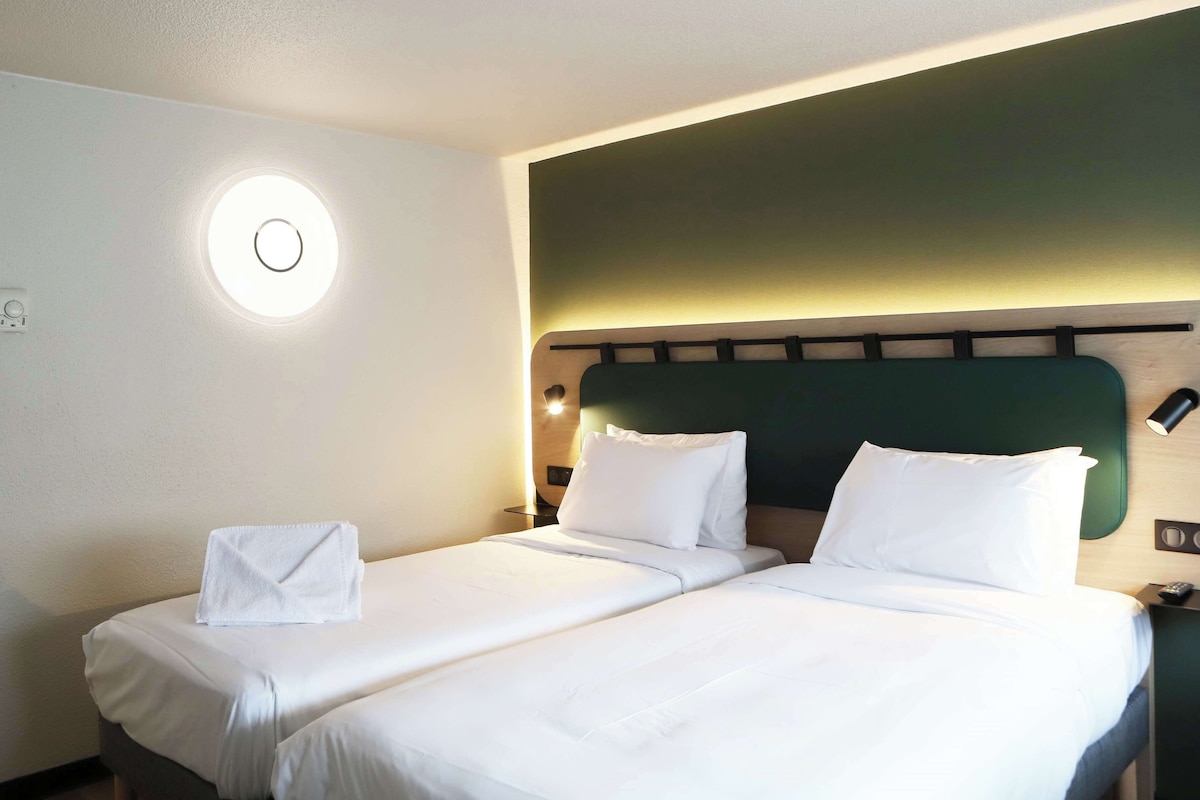 Une chambre d’hôtel comprenant un lit, un applique murale ronde, un thermostat et une table de chevet avec une lampe.