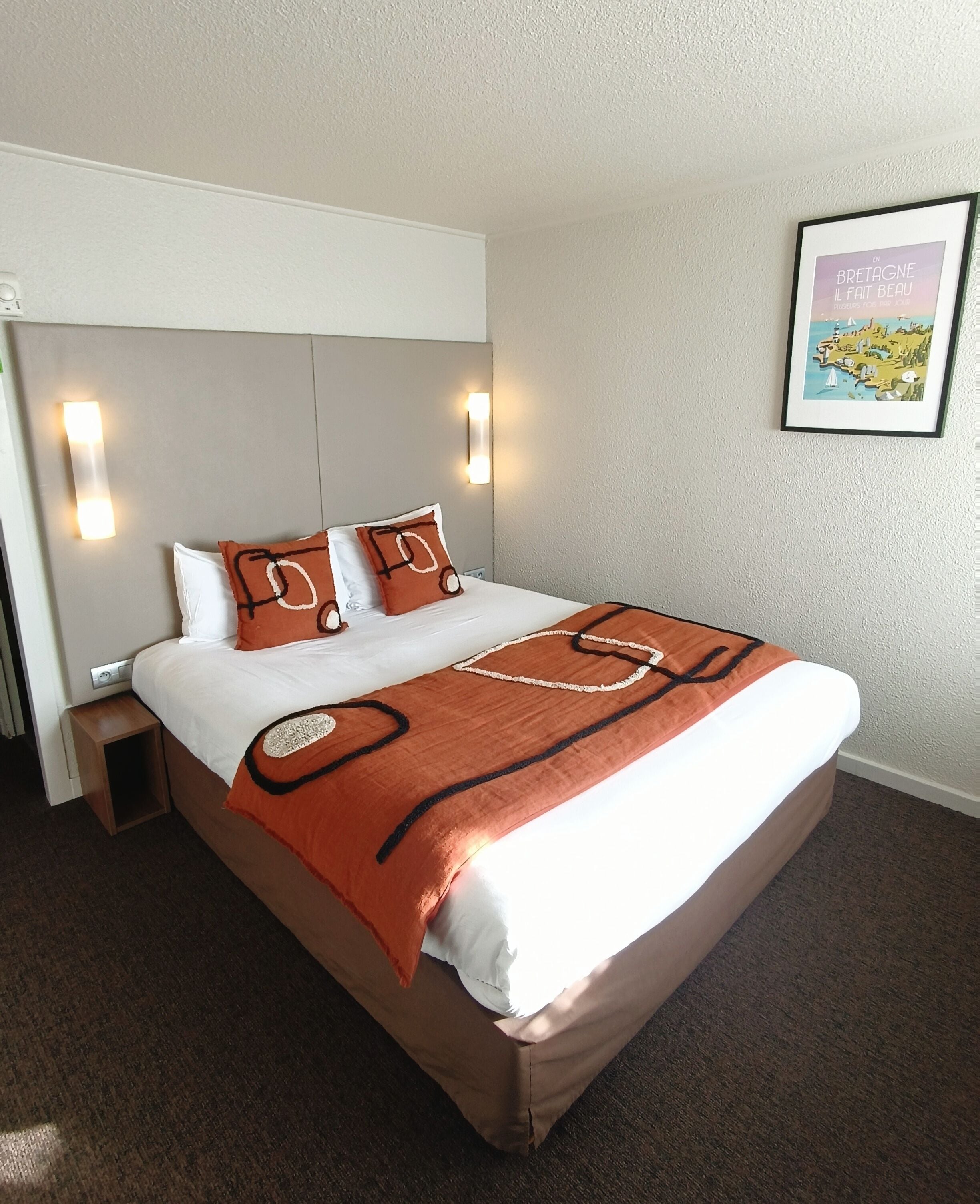 A neatly made bed with a brown blanket and two pillows, a headboard with wall-mounted lights, and a framed map on the wall.