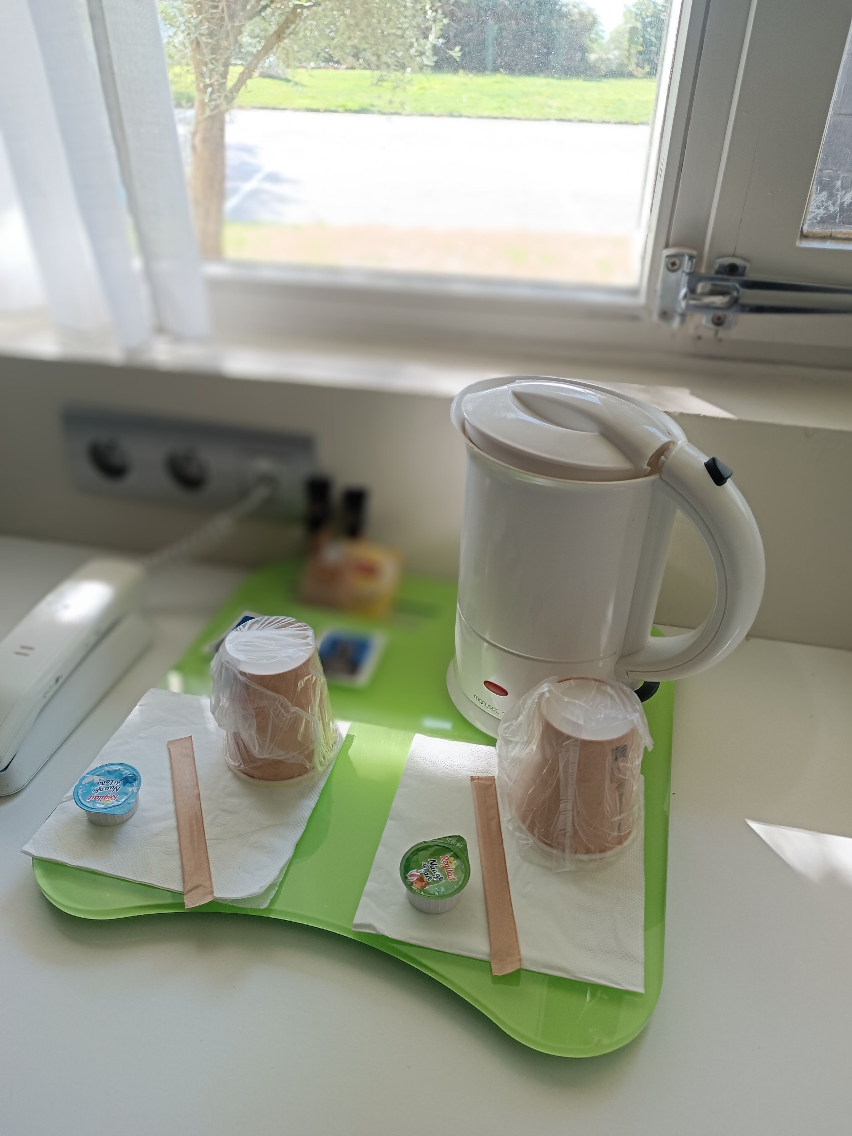 Keittiön työtasolla on kahvinkeitin, vihreä tarjotín, jossa on kaksi pakattua tuotetta, ja ikkuna, josta avautuu näkymä kadulle ja puille.