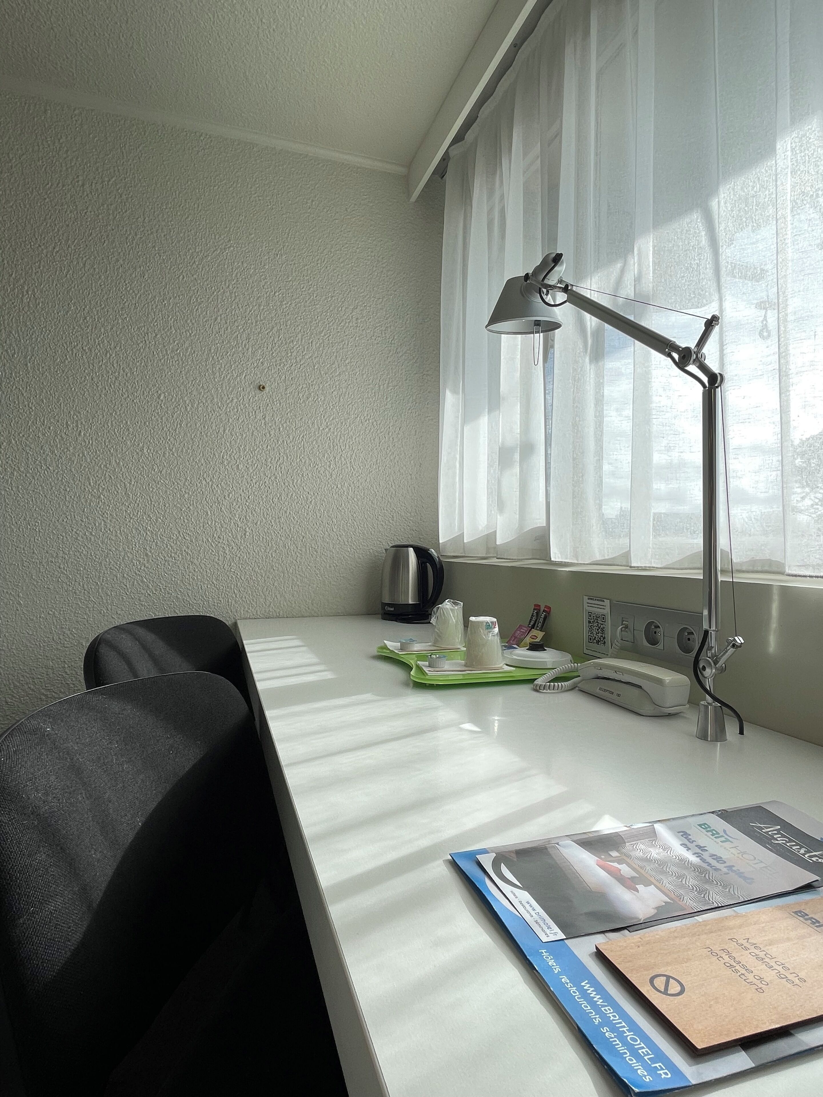 Ein Hotelzimmer mit weißem Schreibtisch, schwarzem Stuhl, Schreibtischlampe, einem Fenster mit durchsichtigen Vorhängen, einem Wasserkocher, einem Telefon und einer Broschüre.