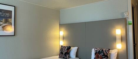 Two single beds with white linens and patterned pillows, a framed picture on the wall, and a light switch on the wall.