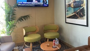 Una sala de estar moderna con un televisor mostrando un noticiero, dos sillones verdes, un sofá de cuero marrón, una mesita redonda con una copa y aperitivos, y un cuadro con una vista de la ciudad.