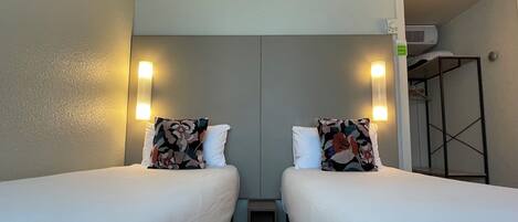 Habitación de hotel con dos camas individuales, cada una con un cojín estampado y uno blanco. Hay luces fijadas a la pared a ambos lados de las camas. Un pequeño mesita de noche está entre las camas, y se observa una estantería metálica en la esquina.