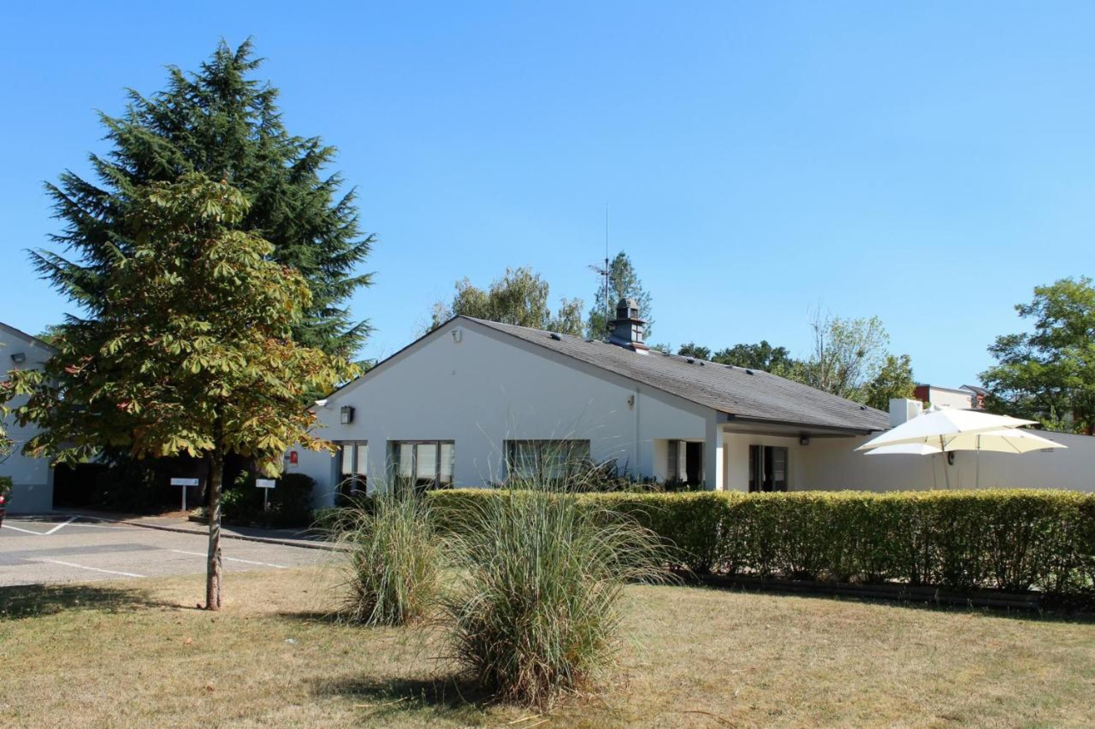 A single-story house with a white exterior, a sloped roof, and a chimney. There is a tree in the front yard and a hedge along the side of the house.