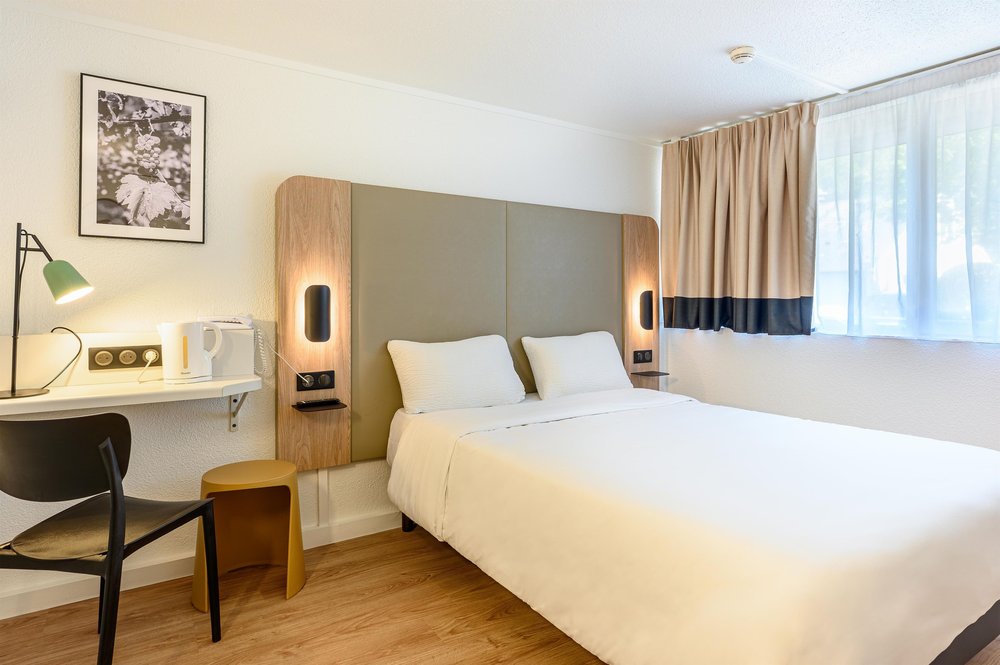 Una habitación de hotel con cama, escritorio con lámpara, silla y un cuadro en la pared.