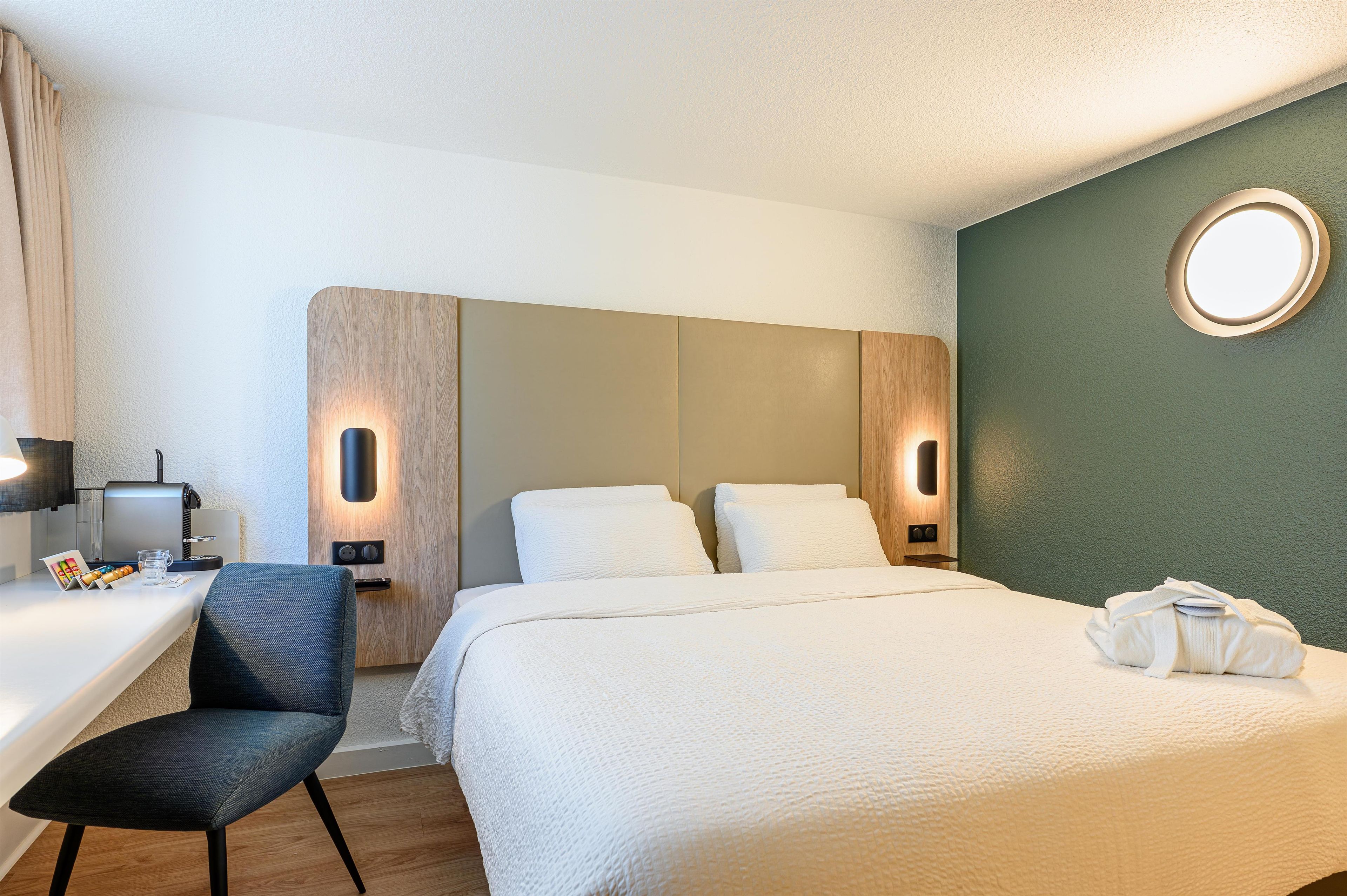 A hotel room with a bed, a desk, a chair, and a round mirror on the wall.