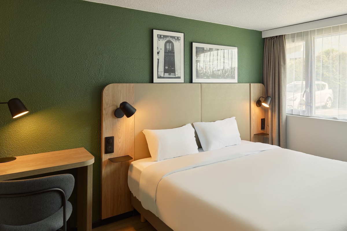 A hotel room with a bed, two bedside tables with lamps, a chair, a window with curtains, and two framed pictures on the wall.